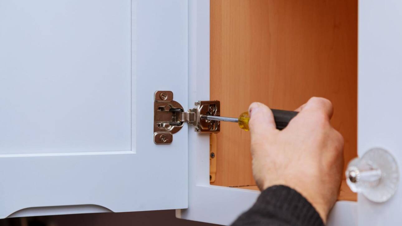 A close-up of a man's hand adjusting a cabinet door hinge on kitchen cabinets.
