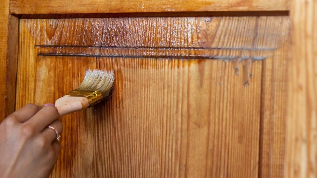 Close-up of a girl's hand staining a wooden kitchen cabinet door with a brush.