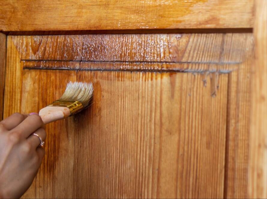 Close-up of a girl's hand staining a wooden kitchen cabinet door with a brush.